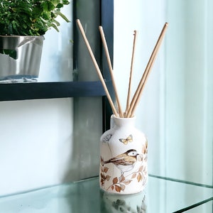 May include: White ceramic diffuser bottle with a bird and butterfly design. The bottle has brown and beige accents and is filled with natural reed diffusers.