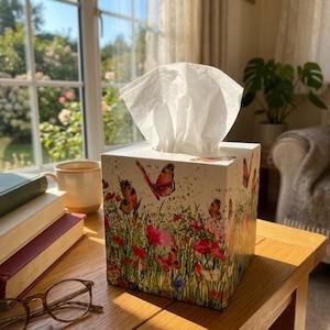 May include: A square tissue box cover featuring a colourful print of orange and black butterflies fluttering over a field of red poppies and green grass. A single white tissue is pulled through the top opening of the box, which sits on a wooden surface.