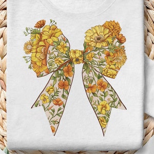 May include: A white t-shirt with a large floral bow design. The bow is made of yellow and orange flowers with green leaves. The bow is centered on the shirt.