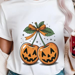 Può includere: T-shirt bianco con un design a tema Halloween. La grafica raffigura due ciliegie arancioni con facce di jack-o'-lantern intagliate, foglie verdi e un fiocco arancione. Piccoli accenti di stelle nere circondano il disegno.