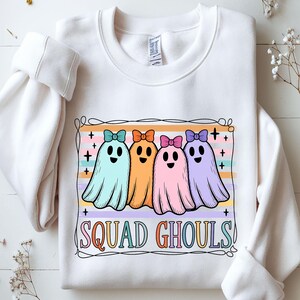 Peut inclure: Sweat-shirt blanc avec un motif de quatre fantômes de dessins animés aux couleurs pastel. Les fantômes portent des nœuds et le texte "SQUAD GHOULS!" est en dessous, dans une police colorée.