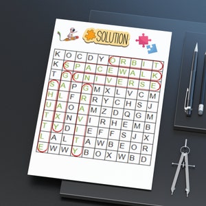 Może przedstawiać: Biała kartka papieru z łamigłówką wyszukiwania słów o tematyce kosmicznej. Szukane słowa są obrysowane na czerwono, ze słowem „SOLUTION” na górze. Ołówki i cyrkiel leżą na ciemnej powierzchni.