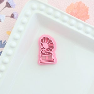 May include: A pink plastic cookie cutter shaped like a vintage record player. The cutter has a detailed design with a horn, a record, and a base.