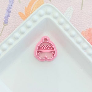 May include: Pink plastic cookie cutter shaped like a winter hat with a pom pom on top. The hat has a diamond pattern on the brim.