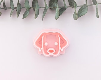 May include: A pink plastic cookie cutter shaped like a dog's head. The cookie cutter has a simple design with a rounded head, two ears, and a nose.