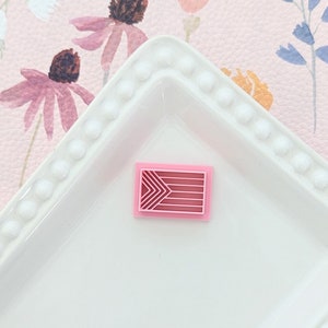 May include: A pink geometric design polymer clay charm with a white background.