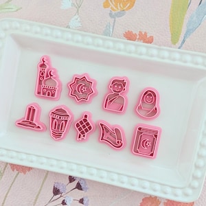 Può includere: Una collezione di formine rosa di varie forme, tra cui una moschea, una stella e una mezzaluna, e figure umane, disposte su un vassoio rettangolare bianco. Il vassoio è posto su una superficie a motivi floreali con fiori rosa e arancioni.