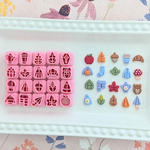 May include: A white rectangular tray holds pink square cookie cutters and colorful fall-themed embellishments. The cutters feature various designs, including a teapot, leaf, and owl. The embellishments include an apple, acorn, and mushroom.