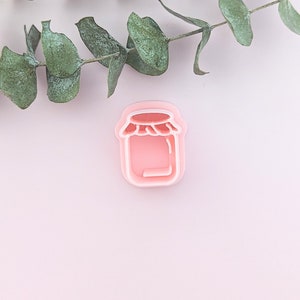 May include: A pink plastic cookie cutter shaped like a jar with a lid. The cookie cutter is on a light pink background.