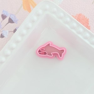 Op de afbeelding: Een roze plastic koekjesvormpje in de vorm van een dolfijn op een wit bord met een decoratieve rand.