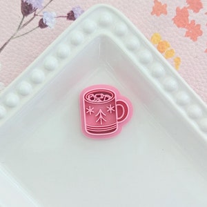 May include: Pink plastic cookie cutter shaped like a mug of hot chocolate with marshmallows and a Christmas tree design. The mug has a white rim and a handle.