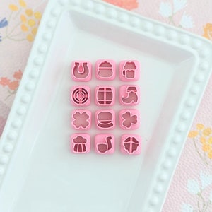 May include: A white ceramic tray with a beaded border, holding 15 pink plastic cookie cutters shaped like various Irish symbols, including a horseshoe, shamrock, beer mug, and leprechaun hat.