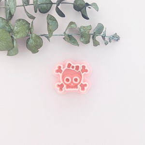 May include: A pink skull-shaped cookie cutter with a bow, set against a white background. The cutter has a layered design with a light pink outline and a darker pink interior. A sprig of green leaves is in the upper left corner.