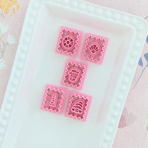 May include: Five pink plastic cookie cutters on a white plate. The cutters are shaped like a seashell, a wheel, a lighthouse, a fish, and a seashell.