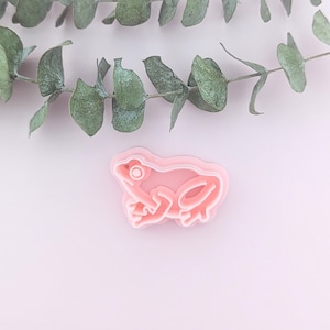 May include: A pink plastic frog cookie cutter on a light pink background with a sprig of eucalyptus leaves.