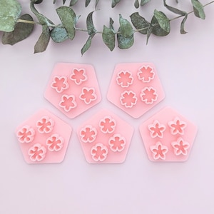 May include: Five pink plastic molds shaped like hexagons with three flower-shaped cutouts in each. The molds are on a light pink background.