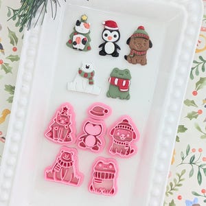 May include: A white rectangular tray displays Christmas-themed resin figurines. The figurines include a Christmas tree, penguin, dog, polar bear, and frog, all wearing festive accessories. Below are pink molds of the same figurines.