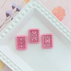 May include: Three pink plastic cookie cutters shaped like tarot cards with different symbols. The symbols are a sun, a moon, and a pentagram.