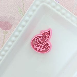May include: A pink fox-shaped cookie cutter with floral and geometric details, on a white rectangular plate with a beaded edge. The background has a floral pattern.