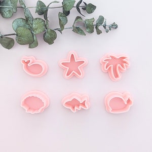 May include: Six pink plastic cookie cutters shaped like a whale, a starfish, a palm tree, a fish, a seashell, and a clam.