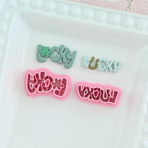 Może przedstawiać: Cztery kolorowe zawieszki z napisami "Lucky" i "Rocka". Zawieszki są w odcieniach różu, zieleni i złota. Zawieszki znajdują się na białej tacy z ozdobnym brzegiem.