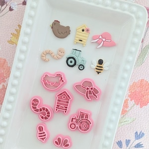May include: A selection of farm-themed cookie cutters and embellishments. The set features a brown chicken, a yellow and brown beehive, a pink sun hat, a light blue tractor, a yellow and black bee, and a pink worm. Additional pink cookie cutters are also included.