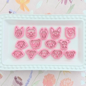 Puede incluir: Un juego de 15 cortadores de galletas de plástico rosa en forma de perro en un plato blanco. Los cortadores están dispuestos en forma de cuadrícula y presentan diferentes razas de perros.