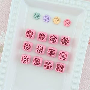 Peut inclure: Un ensemble de tampons artisanaux roses en forme de fleurs et de boutons colorés en forme de fleurs sont disposés sur un plateau rectangulaire blanc. Les tampons présentent divers motifs floraux, et les boutons sont dans des tons de vert, violet, jaune et rose.