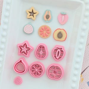 Op de afbeelding: Een verzameling fruitvormige kleibedels en bijpassende roze plastic koekjesvormpjes op een wit dienblad. De set bevat stervrucht-, avocado-, aardbei-, vijg-, citrus- en perzikvormen in pastelkleuren en roze plastic.