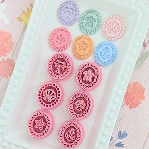 May include: A collection of colorful, round embellishments with coastal designs, including a palm tree, turtle, seashell, coral, starfish, and anchor. The embellishments are in shades of pink, purple, orange, and blue, arranged on a white tray.