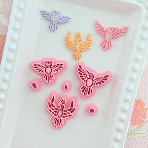 Puede incluir: Una colección de adornos coloridos con forma de pájaro en tonos rosa, morado y naranja. Las piezas decorativas incluyen varios diseños de pájaros, algunos con detalles intrincados en las alas. También hay pequeños adornos redondos.