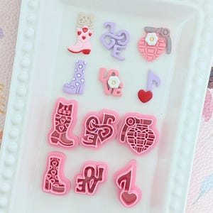 Könnte beinhalten: Eine Sammlung farbenfroher Harzverzierungen in verschiedenen Formen, darunter Stiefel, das Wort "Liebe", eine herzförmige Granate und Noten. Die Farbpalette umfasst Rosa, Lila und Rot, angeordnet auf einem weißen Tablett.