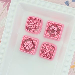 Op de afbeelding: Vier roze vierkante koekjessnijders met verschillende ontwerpen: een vogel, een vrucht, koraal en een geometrische vogel. De snijders zijn gerangschikt op een witte rechthoekige bak. De achtergrond is een lichtroze bloemenpatroon.