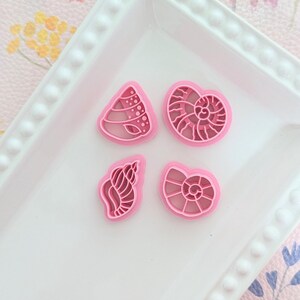 Puede incluir: Cuatro cortadores de galletas rosas con forma de concha marina dispuestos sobre un plato rectangular blanco. Los cortadores presentan diferentes diseños de conchas, incluyendo un nautilus y una caracola. El plato está sobre una superficie con estampado floral rosa.
