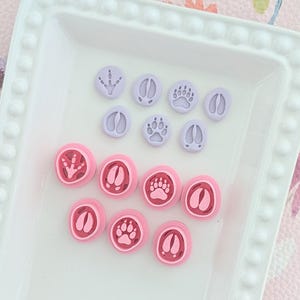 Peut inclure: Un ensemble de petits tampons en argile circulaires présentant diverses empreintes d'animaux. Le lot comprend sept tampons plats violet clair et sept emporte-pièces circulaires roses avec des motifs de pattes et de sabots.