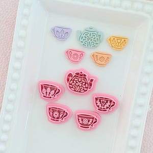 Peut inclure: Une collection d'ornements miniatures sur le thème du goûter. L'ensemble comprend des tasses à thé et des théières roses, jaunes, violettes et turquoise, disposées sur un plateau rectangulaire blanc avec un bord perlé.