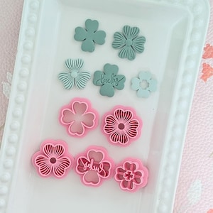 Może przedstawiać: Kolekcja foremek do ciastek i stempli do fondantu w kształcie kwiatów i koniczyny w odcieniach różu i turkusu. Niektóre stemple mają napis "lucky". Przedmioty ułożone są na białej tacy.