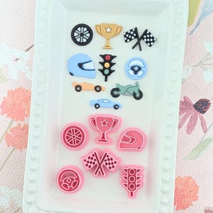May include: A selection of racing-themed embellishments in black, gold, blue, and pink. The collection features a wheel, trophy, checkered flags, helmet, steering wheel, car, motorcycle, and traffic light, presented on a white tray.