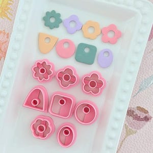 May include: A selection of polymer clay cutters in diverse shapes and colours, including flowers, circles, and geometric forms. The cutters are arranged on a white tray, with shades of pink, green, orange, and purple. These tools are ideal for crafting and jewellery making.