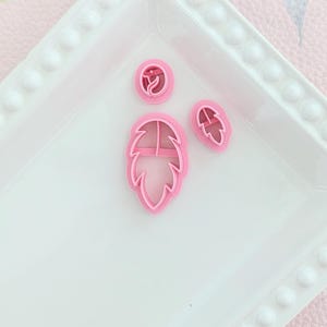 May include: Three pink plastic cookie cutters in the shape of leaves. The cutters are on a white plate with a beaded edge.