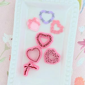 Può includere: Una collezione di abbellimenti a forma di cuore rosa e viola su un vassoio bianco. Gli abbellimenti includono cuori con fiocchi, disegni floreali e motivi a filo spinato. Il vassoio è su una superficie a motivi floreali rosa.