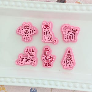May include: A set of six pink cookie cutters in various Halloween-themed shapes, including a bat, cat, deer, goose, and gnome. The cutters are arranged on a white rectangular tray, perfect for baking spooky treats.