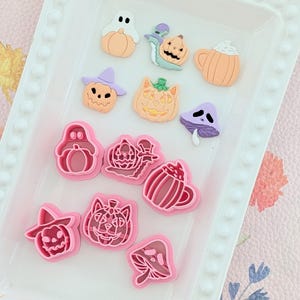 May include: A selection of Halloween-themed resin decorations and cookie cutters. The decorations include a ghost, pumpkins, and a mushroom. The cookie cutters are shaped like a ghost, pumpkins, a mug, and a mushroom. The colours are orange, purple, and pink.