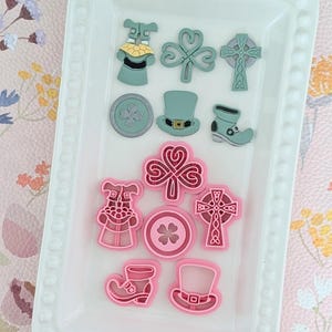May include: A collection of St. Patrick's Day themed cookie cutters in green and pink. The shapes include shamrocks, leprechaun hats, boots, and Celtic crosses, arranged on a white rectangular tray. The tray is on a floral patterned surface.