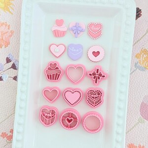 May include: A selection of pink and lilac heart-shaped and cupcake-shaped cookie cutters displayed on a white tray. The cutters feature heart, bow, and "Love" designs, perfect for baking. The tray is white.