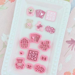 May include: A white rectangular tray containing pink and red decorative embellishments. The embellishments include teddy bears, playing cards with the word "LOVE", hearts, dice, and floral designs. The tray is set against a floral background.