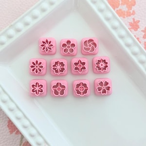 May include: A white rectangular serving dish holds twelve pink flower-shaped cookie cutters. The cutters have various floral designs and are arranged in three rows of four. The background is a pink floral pattern.