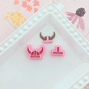 May include: Three pink plastic cookie cutters shaped like Viking helmets. The cutters are on a white plate with a beaded edge.