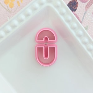 May include: A pink plastic cookie cutter in the shape of the letters "CD" sits on a white rectangular plate with a beaded edge. The background is a pink floral pattern.
