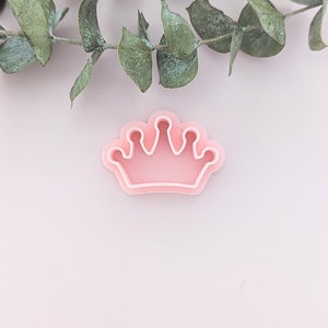 May include: A pink plastic crown cookie cutter. The cutter has a detailed design with five points on the top.